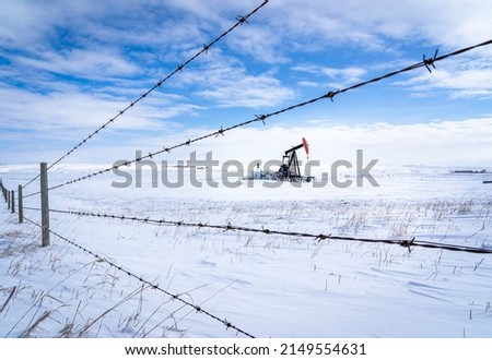 An oil pump jack working in a snow covered agriculture field along a barbed wire fence on the Canadian prairies in Rocky View County Alberta Canada.