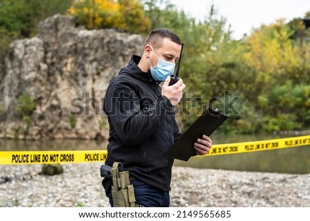 One man police detective or inspector securing crime scene using radio for communication waiting for back up during investigating procedure collecting evidence near the river in day nature copy space Royalty-Free Stock Photo #2149565685