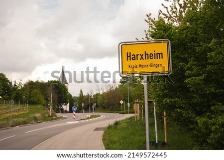 entrance sign of the village Harxheim in Rhineland Platine, Germany with bushes and a church in the background at a cloudy day in spring