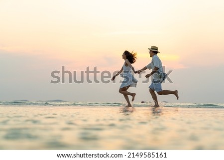 Happy Asian family couple holding hand and walking together on tropical beach at summer sunset on beach vacation. Cheerful husband and wife relax and enjoy outdoor lifestyle on holiday travel vacation