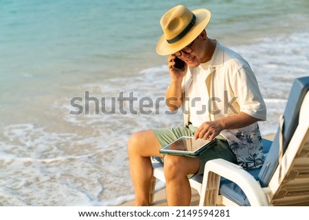 Asian businessman sitting on beach chair working on digital tablet and talking on mobile phone for business stocks trading. Male freelancer enjoy outdoor lifestyle work and travel on summer vacation