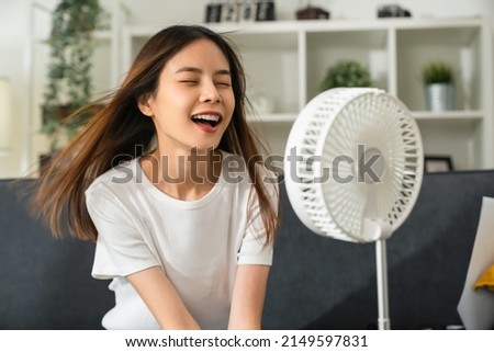 Young smiling asian woman sitting on couch and turned on fan to cool herself with suffers from too hot weather in living room. Royalty-Free Stock Photo #2149597831