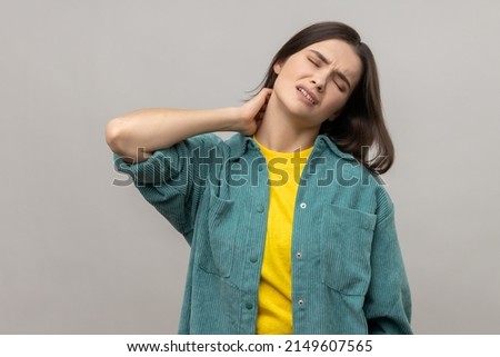 Unhealthy dark haired woman touching neck feeling pain and numbness, worried about muscle tension, osteochondrosis, wearing casual style jacket. Indoor studio shot isolated on gray background.