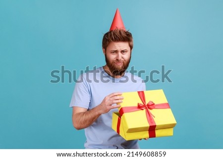 Portrait of sad bearded man in party cone opening gift box and looking at camera with disappointed sad expression, unwrapping bad present. Indoor studio shot isolated on blue background. Royalty-Free Stock Photo #2149608859