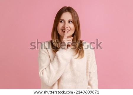 Portrait of optimistic positive young blond woman standing, showing hush sign and looking away, sharing secret, wearing white sweater. Indoor studio shot isolated on pink background.