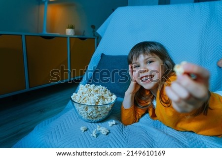 Little girl watching movie at home eating popcorn. Selective focus. Blue light from the screen.