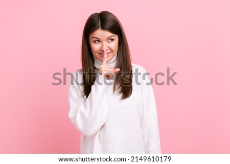 Funny girl keeping finger near mouth, shows secret gesture, asking keep calm, don't tell to anybody, wearing white casual style sweater. Indoor studio shot isolated on pink background.
