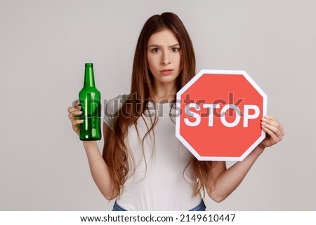 Portrait of anxious beautiful woman showing alcoholic beverage beer bottle and stop sign, warning and worrying, wearing white T-shirt. Indoor studio shot isolated on gray background.