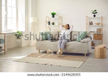 Happy lady leaning back on comfy sofa in cozy bright living room in her own apartment. Relaxed woman enjoying free time on quiet weekend or day off and sitting hands behind head on soft couch at home Royalty-Free Stock Photo #2149628395
