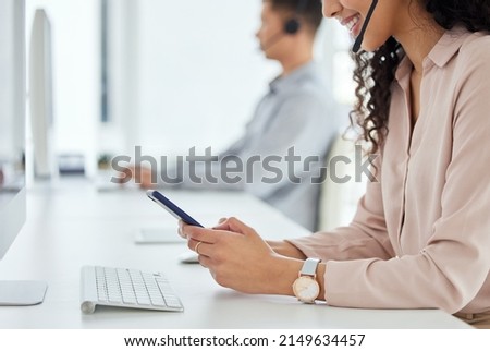 Logging inquiries through multiple digital portals. Closeup shot of a call centre agent using a cellphone while working in an office with her colleague in the background. Royalty-Free Stock Photo #2149634457