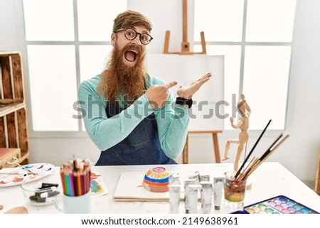 Redhead man with long beard painting clay bowl at art studio amazed and smiling to the camera while presenting with hand and pointing with finger. 