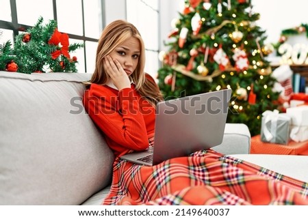 Young caucasian girl using laptop sitting on the sofa by christmas tree thinking looking tired and bored with depression problems with crossed arms. 