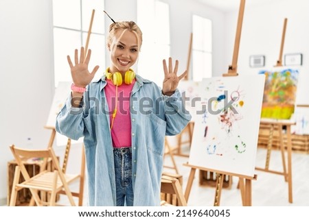 Young caucasian girl at art studio showing and pointing up with fingers number nine while smiling confident and happy. 