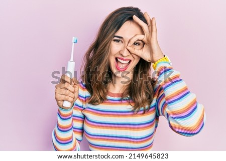 Young hispanic girl holding electric toothbrush smiling happy doing ok sign with hand on eye looking through fingers 