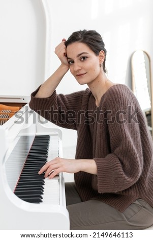 A close-up portrait of a young female musician in elegant casual clothes, playing the grand piano, leaning on it with her elbow