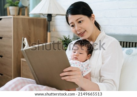 asian mother is reading to her toddler in bed before naptime. lovely baby sitting on mom's legs is looking at picture book. nurturing and lifestyle