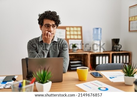 Young hispanic man wearing business style sitting on desk at office looking stressed and nervous with hands on mouth biting nails. anxiety problem.  Royalty-Free Stock Photo #2149676857