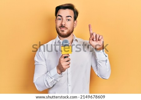 Handsome caucasian man with beard holding reporter microphone surprised with an idea or question pointing finger with happy face, number one  Royalty-Free Stock Photo #2149678609