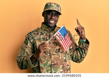 Young african american man wearing army uniform holding american flag smiling with an idea or question pointing finger with happy face, number one 