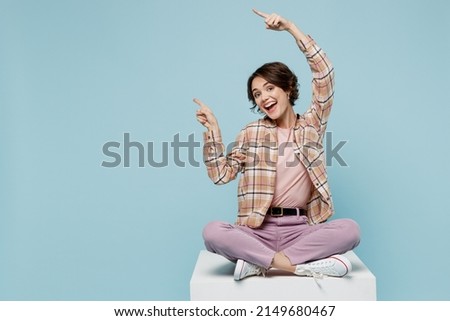 Full size young smiling cheerful happy cool woman 20s wear brown shirt sit on white chair point index finger aside on workspace isolated on pastel plain light blue color background studio portrait.