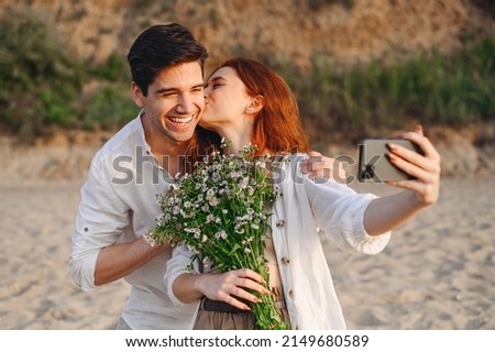 Young couple family man woman in white clothes rest together hold bouquet kiss do selfie shot mobile cell phone post photo social network at sunrise over sea sand beach outdoor seaside in summer day