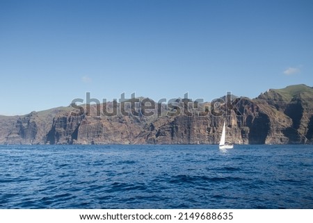 Boat trip for whale and dolphin watching, view of a white sailboat on the horizon, floating in the blue sea and Los Gigantes Cliff in the background. Tenerife, Canary Islands. Spain