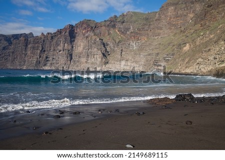 Wave with a lot of foam, breaking on the volcanic black sand beach of the Atlantic Ocean, with a view of the cliff and rocks in the background. Cliff of Los Gigantes, Tenerife, Canary Islands. Spain Royalty-Free Stock Photo #2149689115