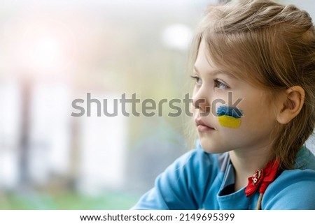 Portrait of an 8-year-old girl with a Ukrainian flag on her face, sitting by the window.