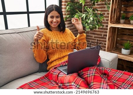 Young hispanic woman using laptop holding virtual currency bitcoin smiling happy and positive, thumb up doing excellent and approval sign 