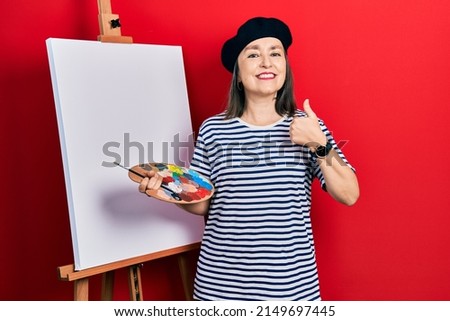 Middle age hispanic woman standing drawing with palette by painter easel stand doing happy thumbs up gesture with hand. approving expression looking at the camera showing success. 