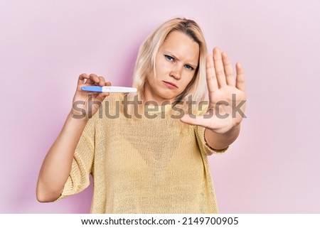 Beautiful caucasian blonde woman holding pregnancy test result with open hand doing stop sign with serious and confident expression, defense gesture 