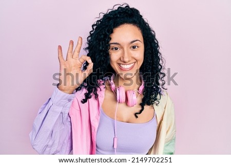 Young hispanic woman with curly hair wearing gym clothes and using headphones smiling positive doing ok sign with hand and fingers. successful expression. 