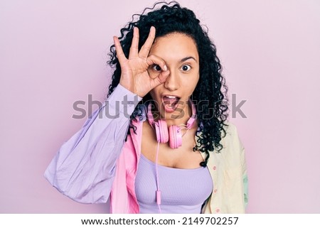 Young hispanic woman with curly hair wearing gym clothes and using headphones doing ok gesture shocked with surprised face, eye looking through fingers. unbelieving expression. 