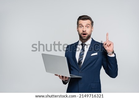 astonished businessman holding laptop and showing idea sign isolated on grey