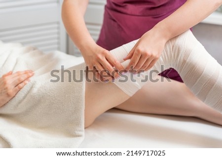 Beautician wraps body of woman in white bandage. Cosmetologist wraps the leg of the customer. Body shaping Royalty-Free Stock Photo #2149717025