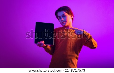 Portrait of boy, child pointing at tablet isolated over purple background in neon light. Watching cartoons online. Concept of emotions, childhood, facial expression. Copy space for ad, design
