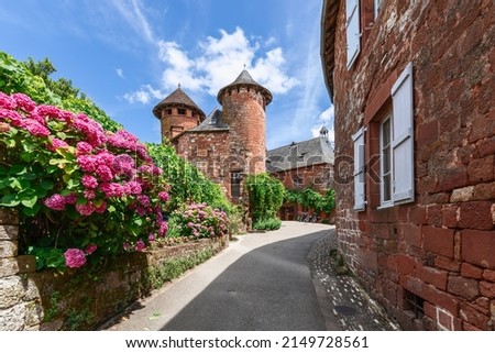 Rhododendrons adorn red sandstone fence along pavement leading to 16 century Ramade de Friac house with two watchtowers of Collonges-la-Rouge village, Correze, New Aquitaine, France Royalty-Free Stock Photo #2149728561