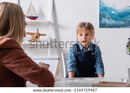 Girl talking near blurred logopedist with clipboard in consulting room Royalty-Free Stock Photo #2149729987