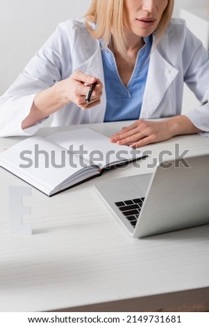 Cropped view of speech therapist holding pen near notebook and laptop in consulting room