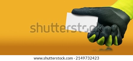 Manual worker with green and black protective work gloves holding a blank business card with copy space, on a yellow and orange background with copy space and reflections.