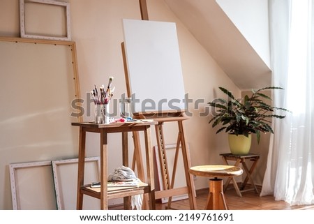 Painting art studio at loft apartment. Empty cozy workplace. Clear canvas on easel and paintbrush on table Royalty-Free Stock Photo #2149761691
