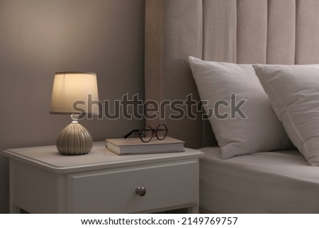 Stylish lamp, book and glasses on bedside table indoors. Bedroom interior elements Royalty-Free Stock Photo #2149769757