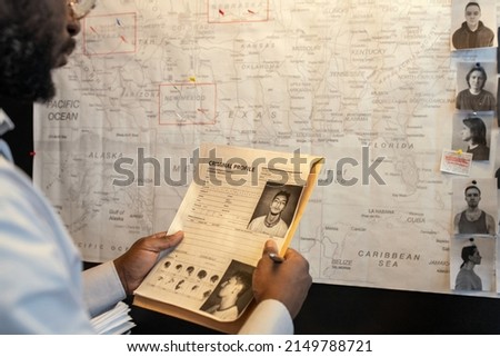 Young African American male FBI agent looking through criminal profile of suspect in front of board with map and photos of suspects Royalty-Free Stock Photo #2149788721