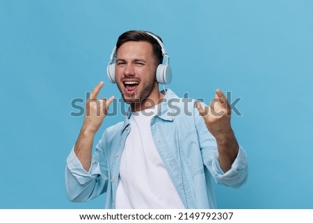 Enjoyed tanned handsome man in casual basic t-shirt headphones listen favorite song show sign of horns rock gesture posing isolated on blue studio background. Copy space Banner Mockup. Music concept