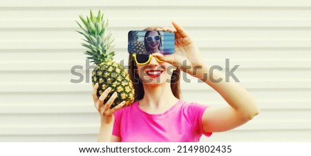 Summer portrait close up of happy smiling woman with pineapple taking selfie by smartphone wearing straw hat, sunglasses on vivid background