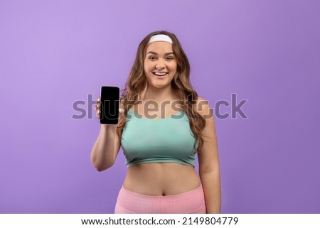 Smiling young european plus size female in sportswear show phone with empty screen, recommends new blog, isolated on purple background. Great offer for workout, sale, app for fitness blog and website