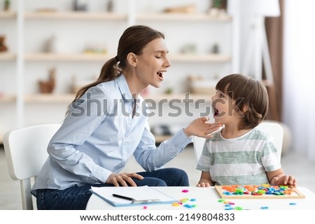 Professional woman speech therapist helping little boy to pronounce right sounds, showing mouth articulation at office, free space Royalty-Free Stock Photo #2149804833