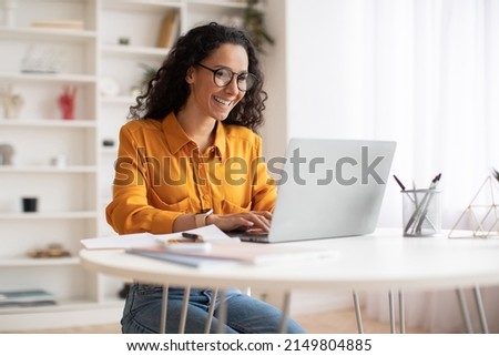 Happy Middle Eastern Businesswoman Using Laptop Typing And Working Online Sitting At Workplace Wearing Eyeglasses. Office Worker Lady Browsing Internet On Computer. Entrepreneurship. Side View