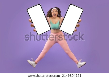 Glad excited young caucasian plus size woman in sportswear jump and freeze in air, show two smartphones with blank screens, isolated on purple background. Health care, app for training, sale and ad
