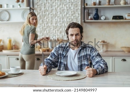 Angry hungry dissatisfied millennial caucasian man with stubble ignoring woman and wait for food in kitchen interior. Quarrel, family problems, stress, social issues and pressure due covid-19 pandemic Royalty-Free Stock Photo #2149804997